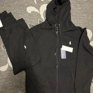 Svart hoodie från Polo Ralph Lauren - Svart hoodie från Polo Ralph Lauren med vit broderad logga på bröstet. Tröjan har huva med snörning, dragkedja framtill och är tillverkad i mjuk bomull. Perfekt för chill dagar och har en clean, stilren look som funkar till allt.