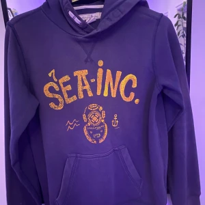 Mörkblå hoodie från H&M  - Snygg mörkblå hoodie med huva från H&M L.O.G.G. i storlek 158/164. Framsidan har ett gult tryck med texten 'SEA-INC.' och en cool dykarmask samt ankare. Tröjan har magficka och långa ärmar, perfekt för chill dagar. 