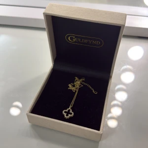 Guldigt halsband med klöver  - Snyggt halsband i guldton med ett klöverformat hänge dekorerat med små glittriga stenar. Kedjan är tunn och elegant, perfekt för att ge din outfit lite extra bling. Levereras i original ask.