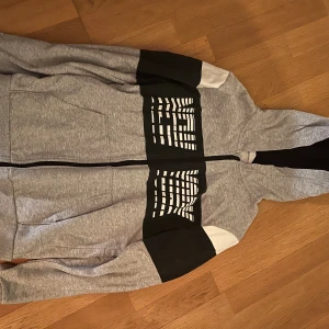 Grå hoodie med svartvitt tryck H&M - Säljer en grå hoodie från H&M med svart och vitt blockmönster över bröstet och ärmarna. Tröjan har huva, dragkedja framtill och stora fickor. Perfekt för chill dagar eller när du vill ha en sportig look.