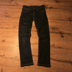 Mörkblå jeans från Nudie Jeans - Snygga mörkblå jeans från Nudie Jeans med klassisk femficksdesign och kontrastsömmar i orange. Raka ben och normal passform, tillverkade i slitstarkt jeanstyg. Bakfickorna har Nudie Jeans signaturbroderi och läderpatch i midjan.