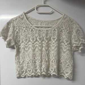 Snygg vit spetsig croppad blus - Superfin croppad blus i vit spets med korta ärmar och rund halsringning. Toppens spets är detaljerad med broderat mönster och har en lätt transparent känsla. Perfekt för sommaren, fest eller festival! 