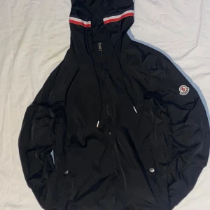 Svart vindjacka från Moncler med huva - Svart vindjacka från Moncler med huva och dragkedja. Jackan har Moncler-logga på ärmen och rödvit rand på huvan. Tillverkad i lätt polyester, perfekt för blåsiga dagar. Snygg och sportig look med praktiska fickor och justerbar huva.