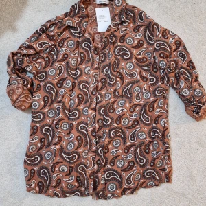 Paisleymönstrad skjorta från Cosy House - Paisleymönstrad skjorta i bruna och beige toner från Cosy House. Skjortan har långärmade ärmar med knappdetalj vid manschetten och klassisk krage. Materialet är mjukt och lätt, perfekt för dig som gillar boho-vibe och snygga mönster. Knappar framtill för enkel styling.