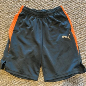 Svarta barn sportshorts från Puma - Svarta sportiga shorts från Puma med orangea sidor och elastisk midja. Klassisk Puma-logga framtill. Perfekta för träning eller chill dagar. Lätta och bekväma i syntetmaterial.