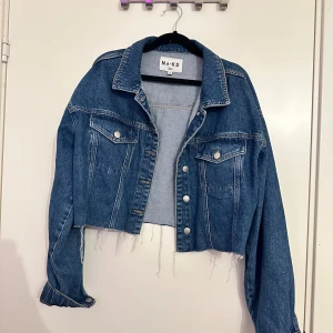 Croppad jeansjacka från NA-KD - Snygg croppad jeansjacka från NA-KD i klassisk blå denim. Jackan har rå kant nedtill, silverfärgade knappar och två bröstfickor med lock. Långärmad modell med avslappnad passform och coola detaljer som fransad nederkant.