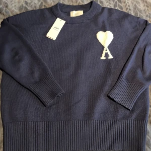 Marinblå stickad tröja från Ami Alexandre Mattiussi  - Snygg marinblå stickad tröja från Ami Alexandre Mattiussi med vit hjärtan-A broderad på bröstet. Tröjan har rund hals, ribbade muddar och lång ärm. Perfekt för dig som gillar stilrena detaljer och vill ha något som sticker ut.