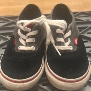 Vans sneakers i svart, grå och röd mocka - Snygga Vans sneakers med svart mocka framtill, grå mocka på sidorna och röd mocka på hälen. Klassisk vit sula med röd rand och vita snören. Vans-logga på sidan och 'Off The Wall' på hälen. Perfekta för en chill streetstyle.