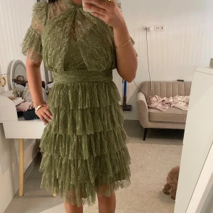 Grön volangklänning i spets - Supersöt grön kortklänning i spets med flera lager volanger och korta ärmar. Klänningen har hög hals, transparenta partier upptill och dragkedja bak. Perfekt för dig som vill sticka ut med en romantisk och trendig look.
