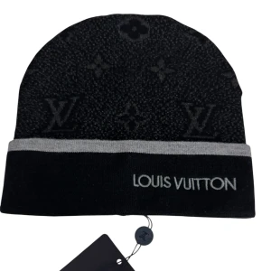 Svart Louis Vuitton mössa med logga - Snygg svart mössa från Louis Vuitton med grå kant och broderad logga framtill. Mössan har diskret LV-monogrammönster över hela ytan och är tillverkad i mjukt stickat material. Perfekt accessoar för att lyfta din vinterstil.