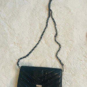 Svart quiltad axelväska Don Donna - Snygg svart axelväska från Don Donna i quiltad design med kedjedetaljer på axelremmen. Väskan är tillverkad i skinnimitation och har flera fack inuti, inklusive ett med dragkedja. Perfekt för att bära det viktigaste med stil.