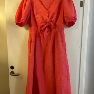 Rosa långklänning med knyt och puffärm - Superfin rosa långklänning med v-ringning, knyt framtill och liten cut-out under bysten. Klänningen har puffärmar och en lätt strukturerad yta i tyget. Perfekt för varma dagar när du vill sticka ut lite extra!