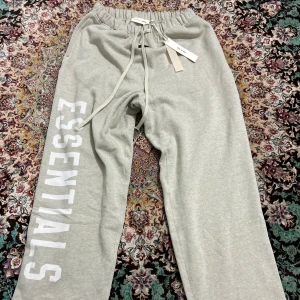 Grå mjukisbyxor Fear of God Essentials - Snygga grå mjukisbyxor från Fear of God Essentials med stor vit logga längs benet. Byxorna har elastisk midja med snörning och är gjorda i mjukt bomullsmaterial. Perfekta för chill eller streetwear och har en relaxed passform.