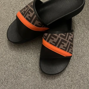 Fendi slides med orange detalj - Snygga slides från Fendi med klassiskt FF-mönster i brunt och svart, samt en fet orange läderdetalj över foten. Skorna har svart platt sula och öppen tå, perfekt för sommaren. Riktigt statement-piece för dig som gillar lyxiga vibes.