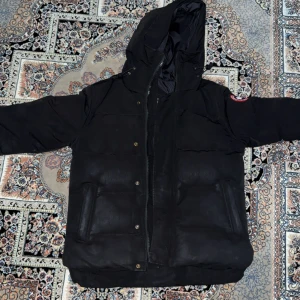 Svart dunjacka från Canada Goose - Svart dunjacka från Canada Goose med huva och klassisk logga på ärmen. Jackan har dragkedja och knappar framtill, två stora fickor och en quiltad design. Perfekt för kalla dagar med sin tjocka och varma fyllning. Snygg och stilren look som funkar till allt.