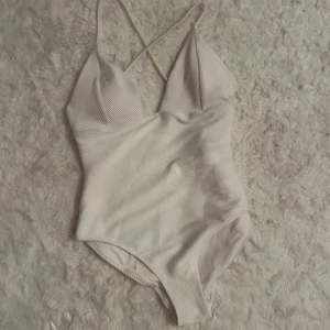 Vit ribbad baddräkt  - Supersnygg vit baddräkt med ribbad struktur och smala axelband som korsas i ryggen och kan sjustera. Baddräkten har v-ringad front och x formad snörning på ryggen. Jag har använt den som en boddy (men den är i baddräktmaterial såklart) den passar S-L