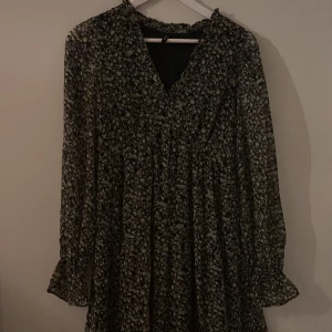 Blommig långärmad klänning Vero Moda - Supersöt svart klänning från Vero Moda med småblommigt mönster i ljusa toner. Klänningen har v-ringning, långa ärmar med volangdetalj vid ärmslut och en luftig, flowy passform. Perfekt för dig som gillar romantiska vibes och vill ha något bekvämt men ändå snyggt.
