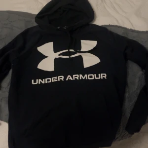 Under armour hoodie - Svart hoodie från Under Armour med stor vit logga och text på bröstet. Tröjan har huva med snörning och är tillverkad i ett mjukt material som passar perfekt för kyligare dagar. Långärmad och loose fit för en avslappnad stil.
