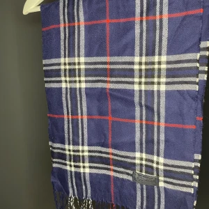 Burberry Halsduk Mörkblå - Snygg halsduk från Burberrys i klassiskt rutig design med marinblå, vit, svart och röda detaljer. Gjord av 100% lammull, mjuk och varm med fransar i ändarna. Perfekt accessoar för att lyfta din stil under kyliga dagar.