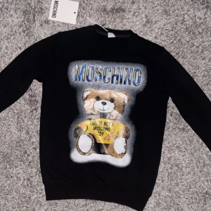 Svart Moschino tröja med björntryck - Cool svart tröja från Moschino med stort färgglatt tryck av en nallebjörn och texten 'This is not a Moschino toy' på framsidan. Tröjan har rund hals, långa ärmar och är tillverkad i mjuk bomull. Perfekt statement piece för dig som gillar streetstyle.