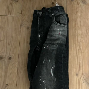Svarta jeans från Dsquared2 med färgstänk - Snygga svarta jeans från Dsquared2 med coola färgstänk och slitningar på både fram- och baksida. Jeansen har klassisk femficksdesign, silverfärgad knapp och Dsquared2-logga vid linningen. Passformen är rak och materialet är robust denim.