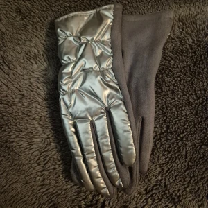 Silvriga quiltade vinterhandskar - Snygga silvriga quiltade handskar med mjuk grå insida. Utsidan har ett glansigt, vadderat material som ger en cool metallic-look, medan insidan är i mjuk fleece. Perfekt för kalla dagar och har en liten blomdetalj på ena handsken.