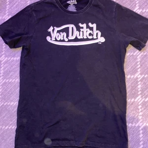 Svart Von Dutch t-shirt  - Cool svart t-shirt från Von Dutch med stort logotryck på framsidan och ett detaljerat grafiskt tryck med vingar och öga på ryggen. Klassisk rund hals och korta ärmar. Materialet är mjuk bomull köptes i USA
