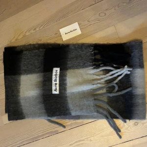 Rutig halsduk från Acne Studios - Snygg och fluffig halsduk från Acne Studios i rutigt mönster med nyanser av svart, grått och vitt. Halsduken har fransar i ändarna och en tydlig Acne Studios-logga. Perfekt accessoar för att lyfta din vinterstil.