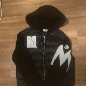 Svart pufferjacka med huva från Moncler - Svart pufferjacka från Moncler med huva och dragkedja framtill. Jackan har ett stort vitt M-tryck på ena ärmen och Moncler-logga. Materialmix med glansig polyester på framsidan och stickade partier. Snygga snören vid huvan och coolt mönster på ärmen.