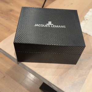 Jacques Lemans silverfärgad klocka - Snygg kronografklocka från Jacques Lemans med silverfärgad urtavla och mesharmband i metall. Medföljer ett brunt läderarmband med krokodilpräglad yta och ett verktyg för att byta band. Klockan levereras i en exklusiv svart box med diskret mönster.