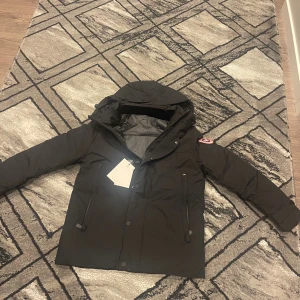  Canada Goose Mcmalian - Helt ny Svart dunjacka från Canada Goose med huva och klassisk logopatch på ärmen. Jackan har dragkedja och knappar framtill, två fickor med dragkedja och en clean, stilren look. Perfekt för kalla dagar och riktigt snygg till streetwear. SITTER SOM STORLEK S