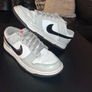Nike Dunk Low light iron ore - Nike Dunk Low sneakers i vitt och ljusgrått med svart Swoosh och röda Nike-detaljer på hälen. Klassisk låg modell med snörning, rund tå och platt sula. Ovandel i skinn och syntet, perfekt för dig som gillar stilrena och tidlösa sneakers. Lite smutsiga och kommer inte med kartong
