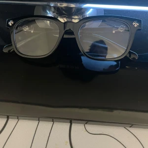 Chrome heart glasögon - Säljer ett par svarta glasögon från Chrome Hearts med rektangulär form och coola silverblommor på bågarna och sidorna. Glasögonen har en chunky vibe och detaljerna i metall ger en lyxig känsla. Perfekt för dig som vill sticka ut med din stil.