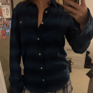 Blå rutig skjorta från Lee - Mörkblå rutig skjorta från Lee med klassisk krage och knappar framtill. Skjortan har lång ärm och en bröstficka med liten logotyp. Perfekt för en avslappnad stil.