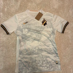 AIK vit fotbollströja Nike limited - AIK fotbollströja från Nike i vit med ljusgrått stadsmotiv och moln på framsidan. Kortärmad med svarta och gula detaljer på ärmsluten, broderat AIK-emblem och guldigt Nike-märke. Materialet är lätt och andas, perfekt för match eller träning.