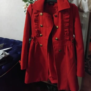 Röd kappa med volang och guldknappar - Snygg röd kappa med volangdetaljer framtill och dubbla rader guldknappar. Jackan har markerad midja, klassisk krage och långa ärmar med dekorativa knappar vid ärmslut. Perfekt för dig som vill sticka ut med en färgstark och trendig look. Jag är 17 år och är ganska liten så passar för de som har storlek s säkert 13-14 158-164 cm