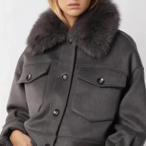 Mörkgrå kappa med päls  - Snygg mörkgrå kappa med stor fluffig pälskrage och stora bröstfickor med knappar. Jackan har en rak passform och stängs med knappar framtill. Perfekt för dig som vill sticka ut med en lyxig vibe. En knapp har åkt av men man får en extra till. Lägger ut för att se vad jag kan få för den för gillar den fortfarande! Finns ej längre 