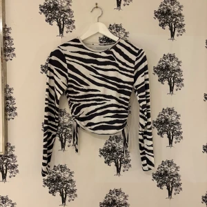 Zebramönstrad långärmad topp med snörning - Säljer en långärmad topp med svartvitt zebramönster och snörning i sidorna för en snygg, croppad look. Toppen har rund halsringning och är tillverkad i ett mjukt, stretchigt material som sitter bekvämt. Perfekt för dig som vill sticka ut med ett djurmönster.