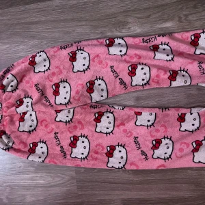Hello Kitty rosa mjukisbyxor - Supergosiga rosa mjukisbyxor med Hello Kitty-tryck över hela byxan. Byxorna har elastisk midja och är gjorda i mjukt fleece-material, perfekta för chill hemma. Söt design med röda rosetter och små hjärtan som detaljer.