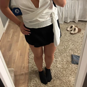 Svart omlottkortkjol med dragkedja - Säljer en svart shorts/kjol med omlott-design och dragkedja i sidan. Kjolen har en stilren look och är lätt att matcha med olika toppar. Materialet känns mjukt och har en lätt struktur, perfekt för dig som gillar minimalistisk stil. Finns ett hål nedanför dragkedjan, ingenting som syns från håll.