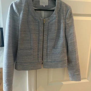 Randig ljusblå kavaj från Stockh LM - Snygg ljusblå kavaj från Stockh LM med diskret randigt mönster och croppad passform. Jackan har rundad hals, två fickor fram och stängs med dragkedja. Perfekt för dig som gillar stilrena detaljer och modern look.