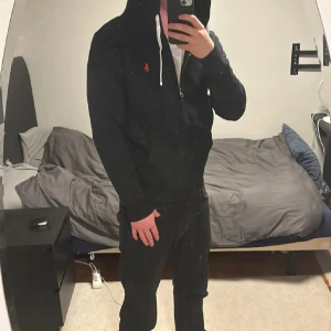 Svart hoodie från Polo Ralph Lauren - Svart hoodie från Polo Ralph Lauren med dragkedja och vit snörning i huvan. Klassisk röd logga broderad på bröstet. Tröjan har långärmad passform och är tillverkad i mjuk bomull, perfekt för chill dagar. Snygg och enkel att matcha med jeans eller joggers.