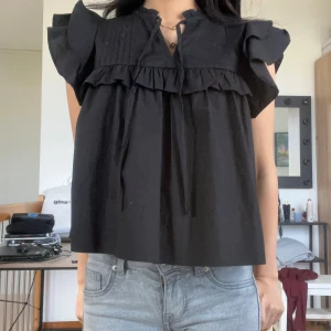 Svart blus med volang från Gina Tricot - Supersöt svart blus från Gina Tricot i storlek XS. Blusen har volangdetaljer vid axlarna och över bröstet, samt knytning vid halsen. Den är ärmlös med breda axelpartier och har en luftig, loose passform. Perfekt för dig som gillar romantiska detaljer och stilren look.