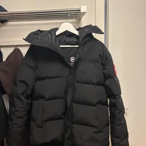 MacMillan Canada goose - Säljer en svart dunjacka från Canada Goose med klassisk logga på ärmen. Jackan har huva, dragkedja och två stora fickor framtill. Perfekt för kalla dagar och har en snygg, puffig look som är riktigt trendig just nu. Andvänd 1 gång