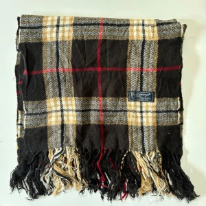 Rutig halsduk från Burberry - Snygg rutig halsduk från Burberry, tillverkad i 100% lammull. Färgerna är svart, beige, vit och röd med klassiskt rutmönster och fransar i ändarna. 
