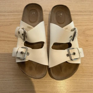 Vita sandaler med dubbla spännen - Säljer ett par vita sandaler med två breda remmar och silvriga metallspännen. Sandalerna har en platt sula och är tillverkade i skinnliknande material med struktur. Perfekta för varma dagar och enkel att matcha med olika outfits.