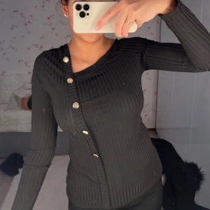 Svart ribbad topp med knappar - Snygg svart ribbad topp med asymmetrisk design och knappar längs ena sidan. Toppen har långa ärmar och en unik offshoulder-stil som ger en trendig look. 