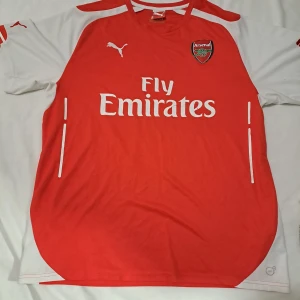 Arsenal Puma  XL  - Arsenal fotbollströja från Puma i rött och vitt med klubbmärke och Fly Emirates-tryck på bröstet. Klassisk design med rund hals och korta ärmar. Tillverkad i lätt och ventilerande polyester, perfekt för match eller träning.