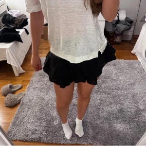 Svart volangkjol i kort modell med inbyggda shorts  - Supersöt svart kortkjol med flera lager volanger som ger en fluffig och lekfull look. Kjolen har en flowy passform och är perfekt för dig som gillar att sticka ut. Passar till både sneakers och boots för en cool vibe. Kjolen har inbyggda shorts. Den är knappt använd så den är i väldigt fint skick. 
