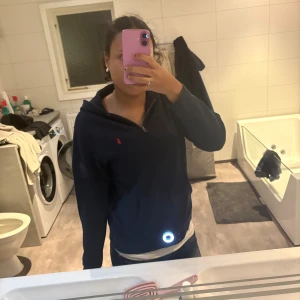 Mörkblå hoodie från Ralph Lauren - Säljer en mörkblå hoodie från Ralph Lauren med röd logga på bröstet. Tröjan har dragkedja framtill, huva och långa ärmar. Perfekt för dig som gillar klassisk streetstyle och vill ha något bekvämt och snyggt.